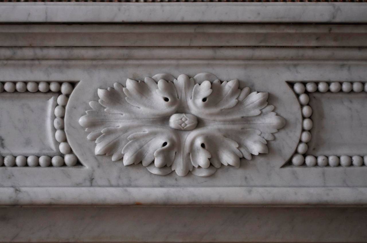 This antique Louis XVI style fireplace was sculpted in Carrara marble during the late 19th century, in Paris. The fireplace is carved with acanthus leaves and pearls. 
The fireplace is sold with its original silvered cast iron insert and its