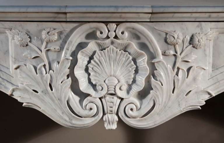 This antique Louis XV style fireplace was made out of a white Carrara marble in the 19th century. The sculpture decoration is opulent. The frieze is curved and ornated in its center of a double shell with foliages and flowers. Jambs features