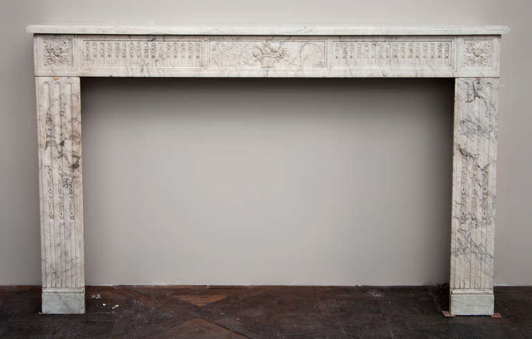 This beautiful antique Louis XVI period fireplace was made out of Carrara marble during the 18th century. Fluted, the fireze is carved in with a fruits basket. Both jambs, with straight lines, are fluted are sculpted with asparagus heads. 
The top