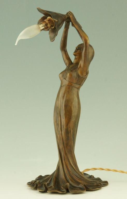An Art Nouveau bronze figural lamp of a lady holding a flower.
By Taupin d’Auge. 
Foundry: Fonderie de Vincennes éditeur. 

Rewired for European electricity.

Fedex shipping: $ 225.