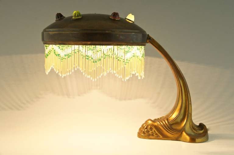 An art nouveau bronze, brass and glass piano or desk lamp.
Ca. 1900.			

Material: 
Patinated bronze, brass shade with glass inlay.   
White, yellow and green frills. 
 			
Size:		
H. 11.4 inch x L.15 inch. x W. 9.8 inch. 	 
H. 29 cm  x L.