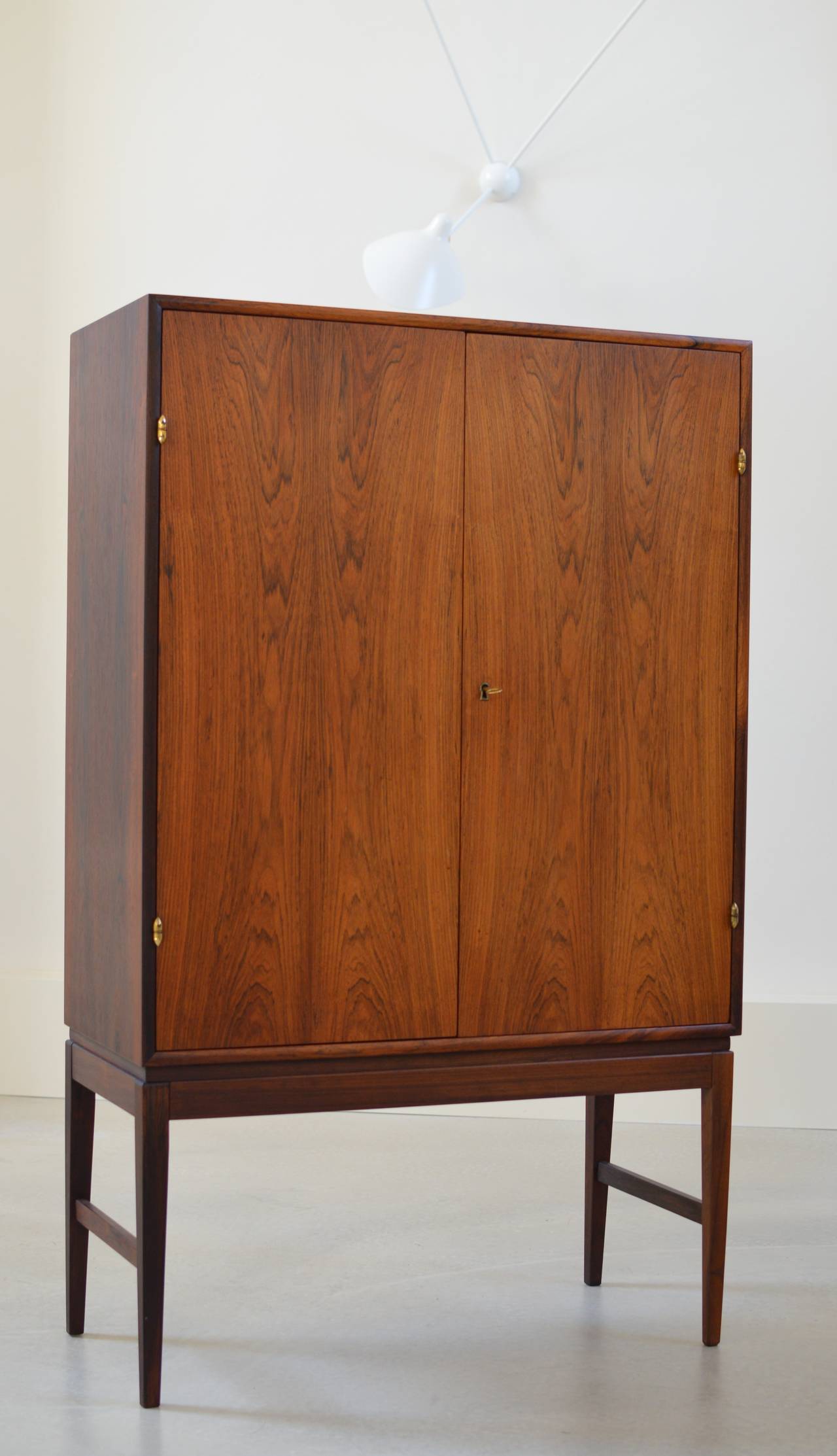 Beautifully crafted refined 1960's Danish Rosewood cabinet with facet cut mirror, shelves and a smaller drawer compartment on high legs legs and remarkably bullet shaped brass hinges. 
The interior shelves are adjustable from which the top shelf is