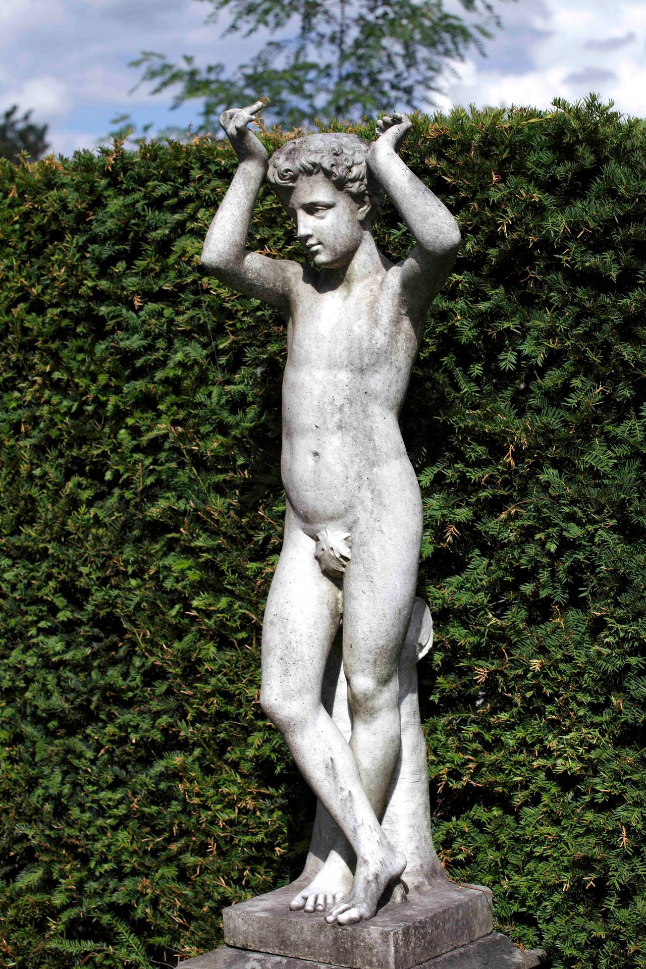 Portrayed standing and nude, his hands raised above his tousled hair, his legs crossed with his weight on the left leg, against a supporting tree stump, on a rectangular section socle, measuring 136 cm. high.