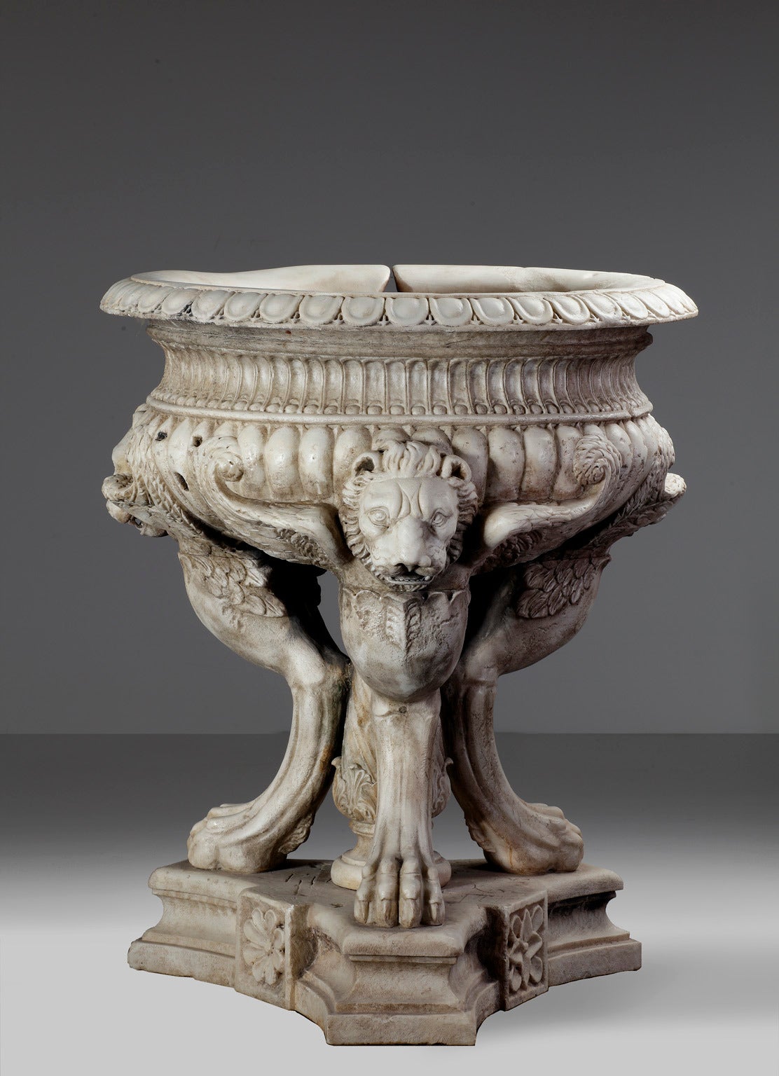 The everted rim above a waisted and fluted band, the lobed underside supported on the spread wings of three adorsed monopodial lions, around a central spiral carved supporting column, the triform base with waisted sides.

The planter is almost