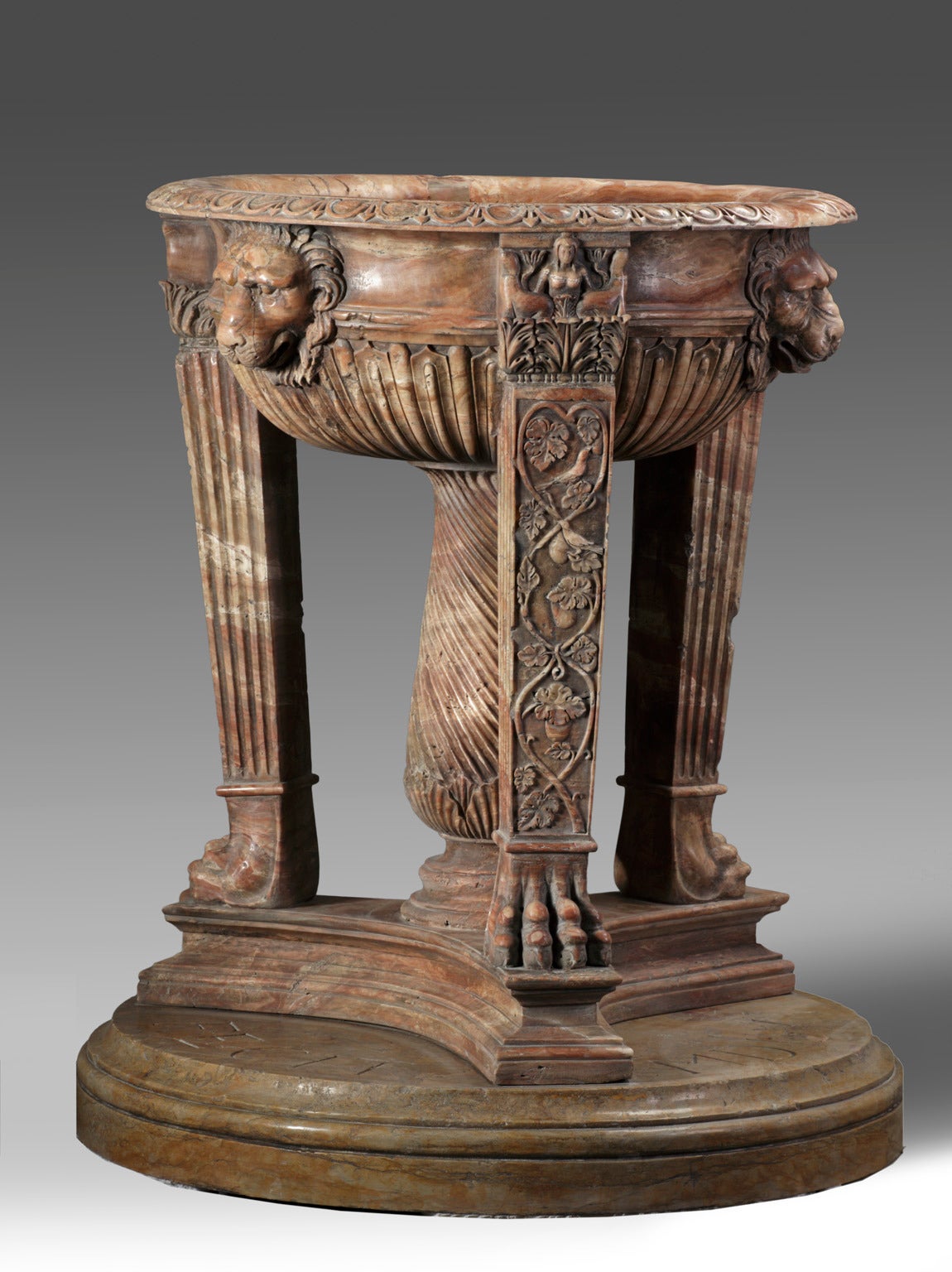 The basin with everted rim above a waisted band with three lion's masks carved in relief, with fluted underside, held on three rectangular section fluted and foliate cast legs with paw feet, around a central spiral carved baluster supporting column,