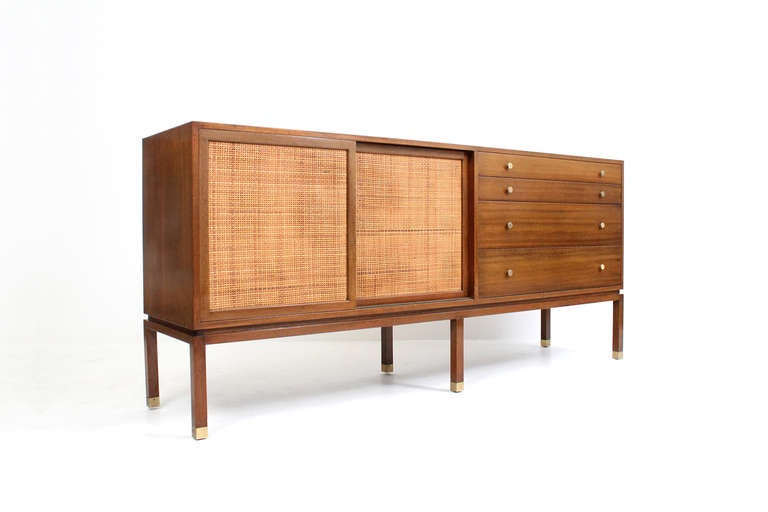 Elegant Harvey Probber credenza with brass details and caned doors.  Cabinet features caned sliding doors on the left revealing a shelf and cork lined drawers for storage and a bank of drawers with solid brass pulls on the right.  Well proportioned