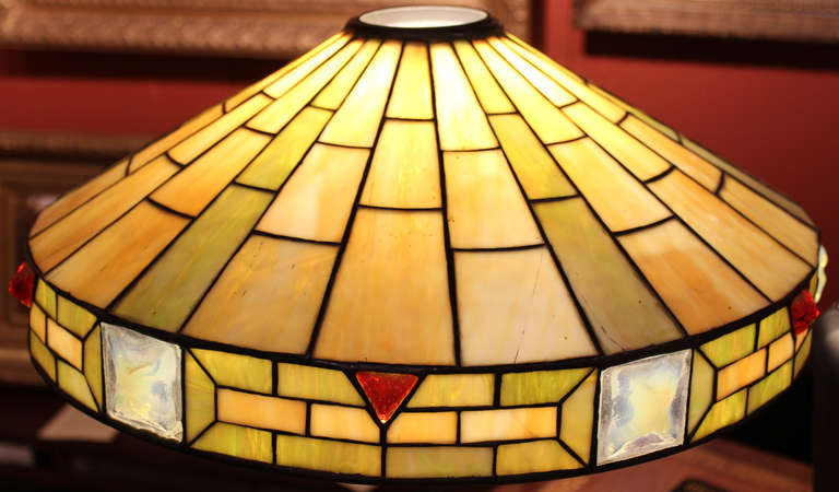This Arts & Crafts lamp with leaded glass shade has subtle greens and yellows, highlighted with a jewels around the outer edge. Originally a gas lamp converted to electric. Stamped on base “505”.