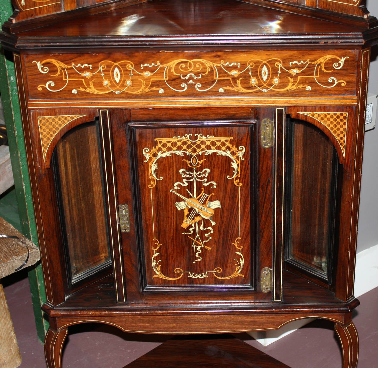 A beautiful two part rosewood inlaid corner cupboard with marquetry, shaped beveled mirrors, broken arched pediment, and single lower door for storage with  inlaid viola and lyre. Makers label on verso “James Shoolbred & Co, Tottenham House,