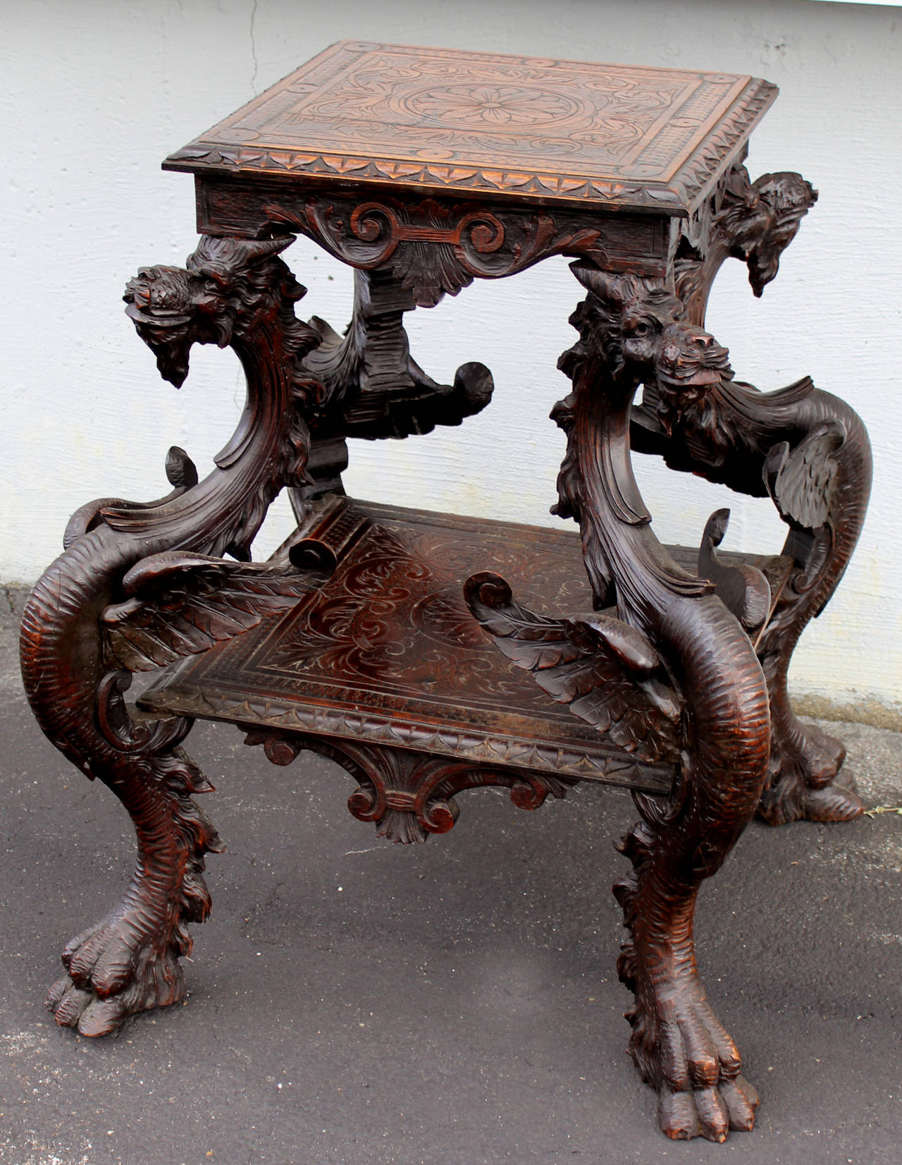 A boldly carved walnut two-tier pedestal table with four carved winged dragons supporting the squared top with foliate carving and conforming medial shelf. Dragon supports terminating in doubled paw feet.