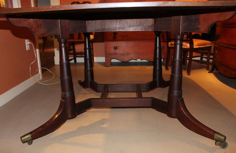This beautiful English mahogany Cumberland action dining table has a rectangular top with rounded corners supported by turned gateleg pedestals. Each leg has its original brass caps and castors.
