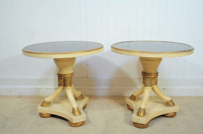 Unique pair of Carved Wood French Regency / Neoclassical Style Round Figural Low End Tables in the manner of Maison Jansen. This stunning pair features a cream painted and gold gilt finish with round antiqued black glass tops. The tripod goat / rams