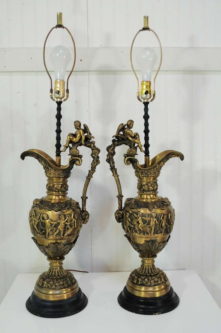 Stately pair of early 20th century bronze ewer figural table lamps in the French neoclassical / classical form. Item features seated winged cherubs on the handles which end at ram’s heads. The lamp bodies feature wonderfully detailed scenes of