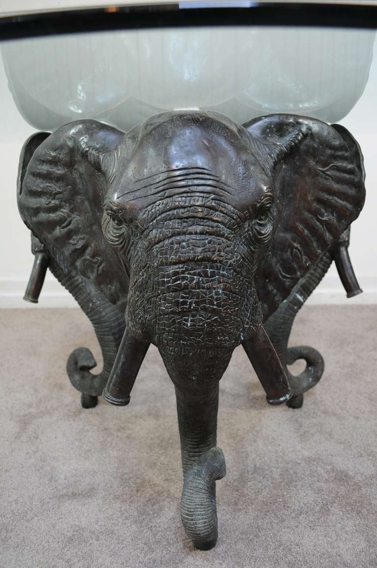 Stunning figural table constructed of a heavy patinated cast bronze triple elephant pedestal base and a round glass top. This item features exceptionally life-like features in the elephant faces and a great patinated and antiqued bronze finish.