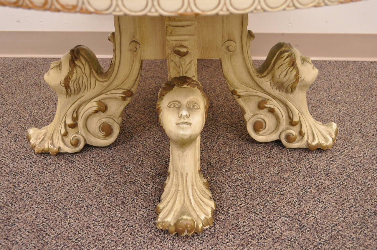 Uniquely carved vintage Hollywood Regency coffee table in the French taste. This elegant item features an inset glass top with white border and gold leaf decorated central pattern. The base is solid carved wood with four female faces and