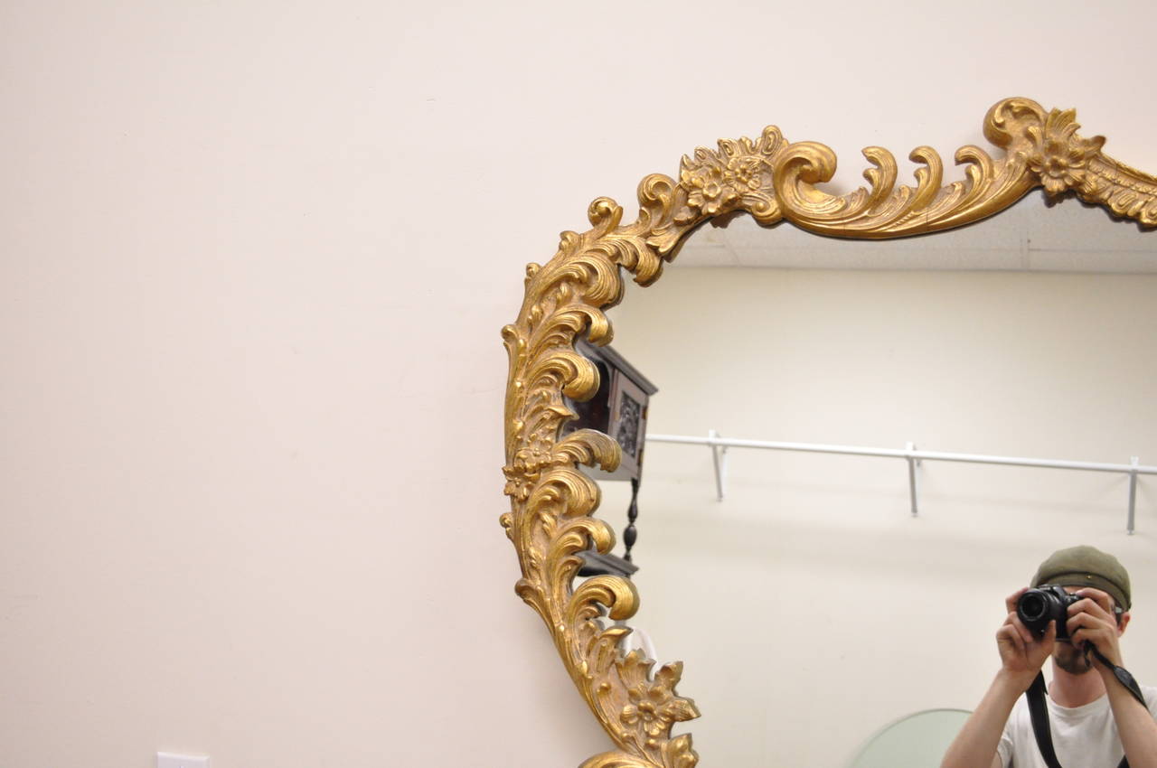 Very ornate and stately French Rococo style wall mirror comprised of gesso over wood. The item features exquisite floral and acanthus motifs, and a brilliant gold gilt finish. A very impressive and eye-catching statement piece. Please note that the