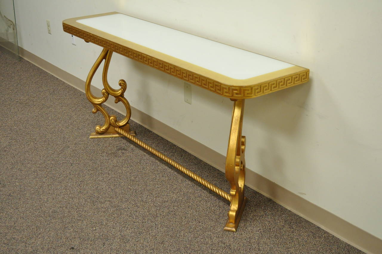 Attractive inset glass top console table with Greek key detailing adorning the apron. The top features a white underlay with gold leaf banding. The base consists of a scrolling stretcher which supports the scroll form shapely legs. Depending on the