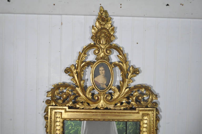 Stunning 1920s Carved Wood and Gold Gilt Italian Wall Mirror with female cameo at the crest in the French Rococo taste. This fine mirror features a ornately acanthus carved wooden frame, and a print of a female portrait at the crest. Made in Italy