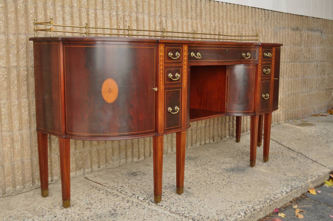 Remarkable, High Quality, English George III Style Serpentine Mahogany Sideboard, Circa Early 20th Century. This fine and substantial piece features a solid brass gallery, satinwood pinwheel, bell flower, and pencil inlay, brass capped feet, hand