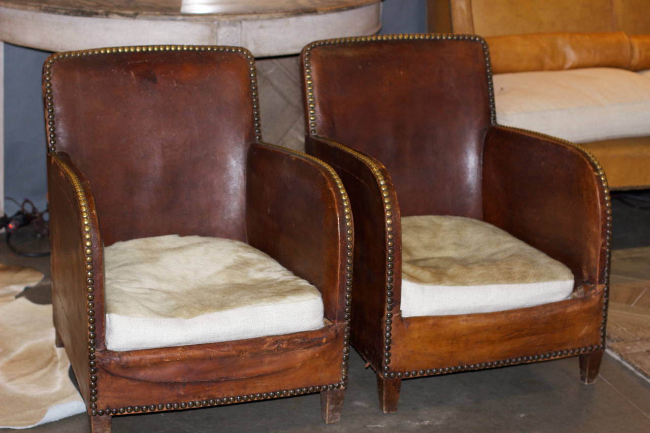 Lovely pair of 1920s French leather club chairs with original brass nailhead trim. Cushions have been recovered with white cowhide and linen. Very comfortable and wonderful patina.