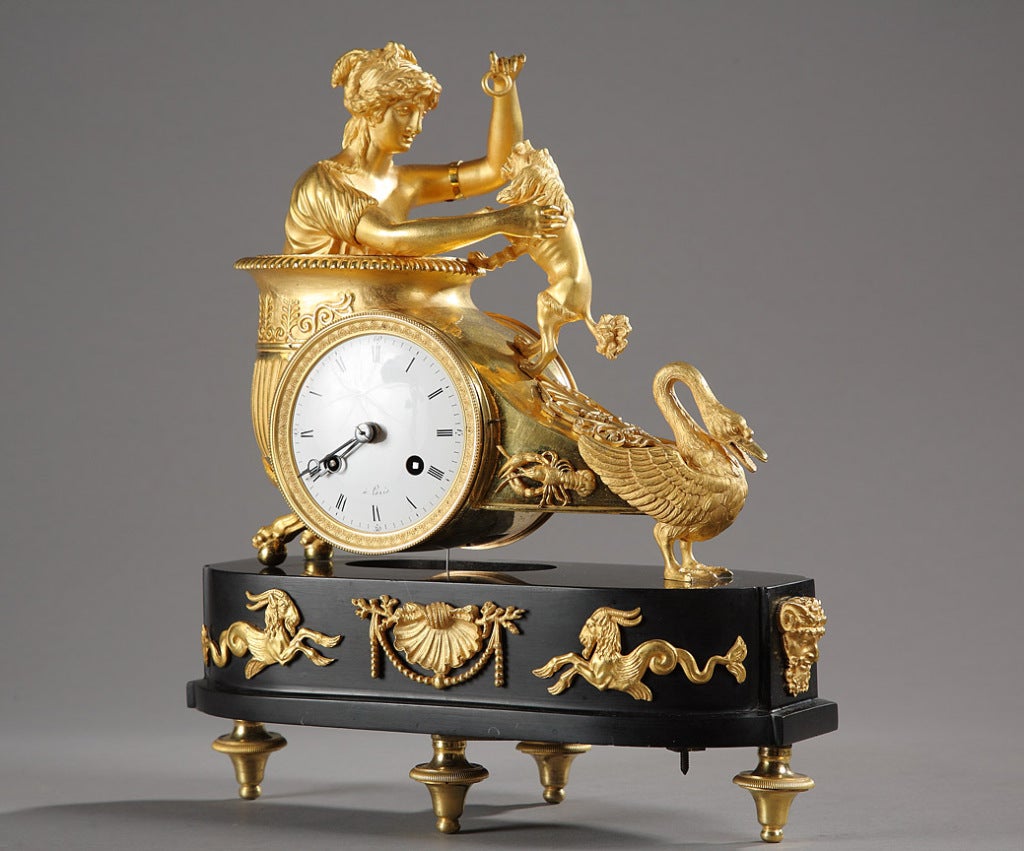 French Empire mantle clock representing a woman draped in antique dress, in a chariot pulled by a swan, playing with a dog. Lobster patterns adorn the sides of the chariot. Oval black marble base, with cut sides resting on four feet. Gilded bronze