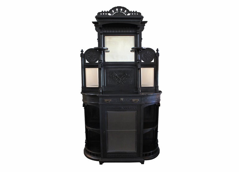large ebonized victorian cabinet with bonnet top, beveled mirrors, and various display shelves. intricate carvings throughout with flamed torch design on the center panel. lower cabinet with beveled glass door and working skeleton key. original