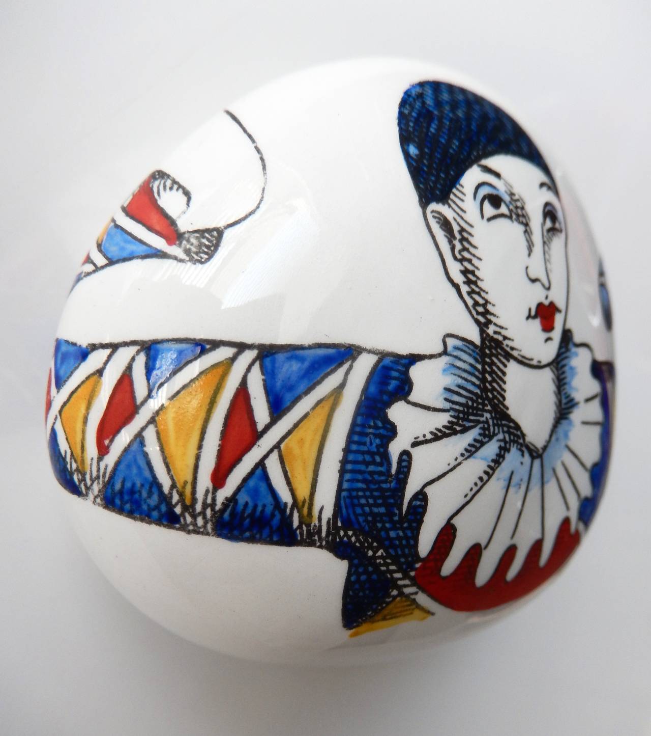 An oval shaped, porcelain paperweight by Piero Fornasetti of a Harlequin or French Pierrot figure. Commedia dell'arte characters are a popular subject in Fornasetti's work. Marked. Beautiful, brightly-colored graphic design. 

(Similar