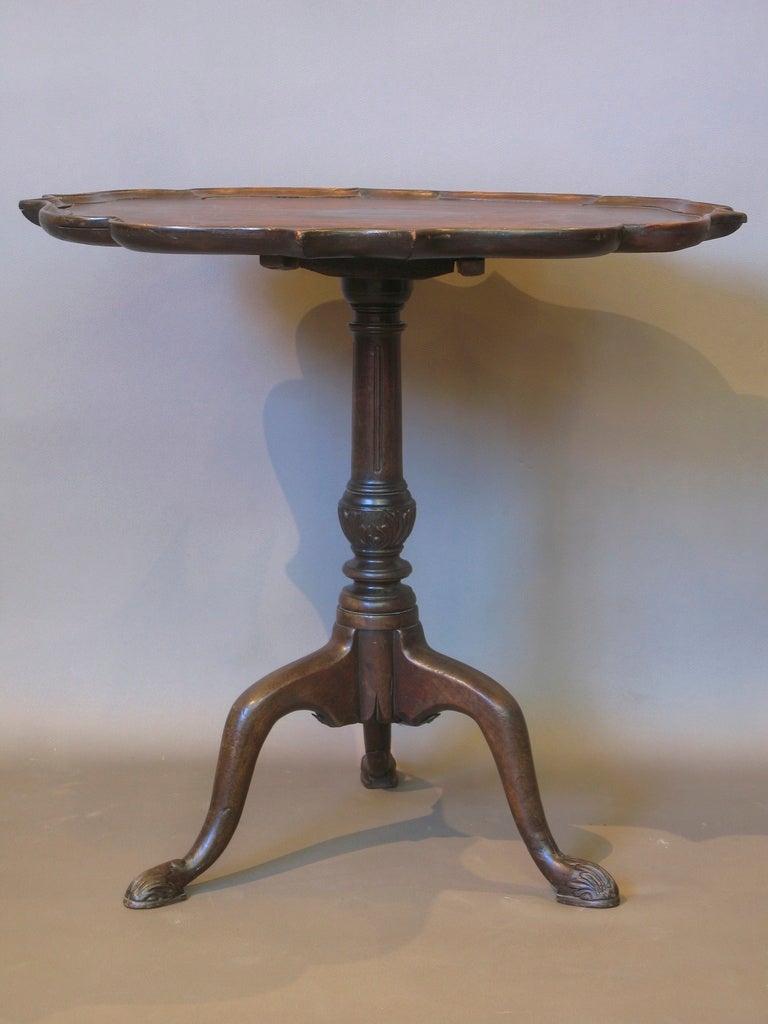 Elegant tea table with an unusually shaped top, having a raised and scalloped carved edge, resting on a baluster column carved with foliate design and supported by three cabriole legs that end in slipper feet, also adorned with carved scrolling