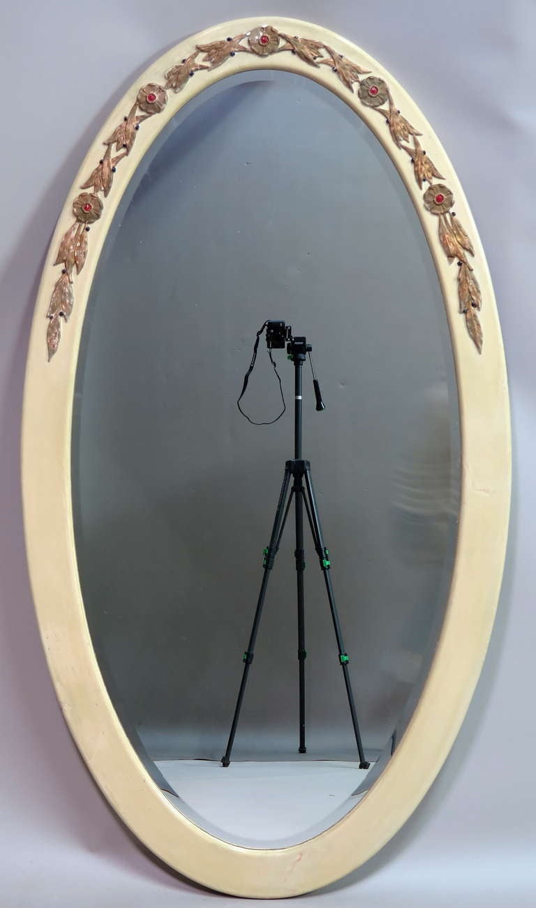 Lovely pair of large French Art Deco oval mirrors, with beveled glass. The wooden frames are carved with a top wreath of flowers and laurel leaves painted gold, red and black on an off-white background.

Very elegant.