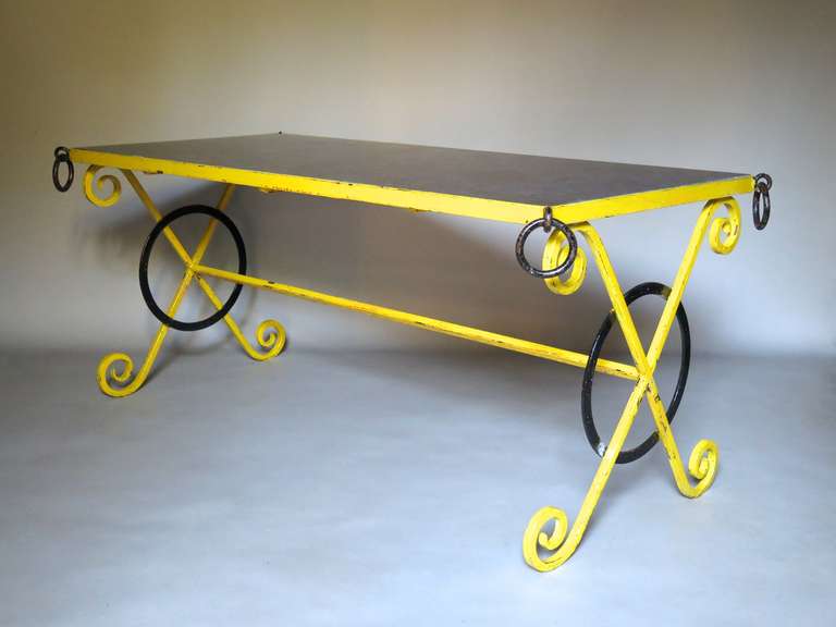 A lovely and colourful outdoor dining table with a wrought iron structure, painted glossy yellow and black (traces of orange beneath). Four wrought-iron rings adorn the corners of the table top which is fitted with a black stone slab.
Although most