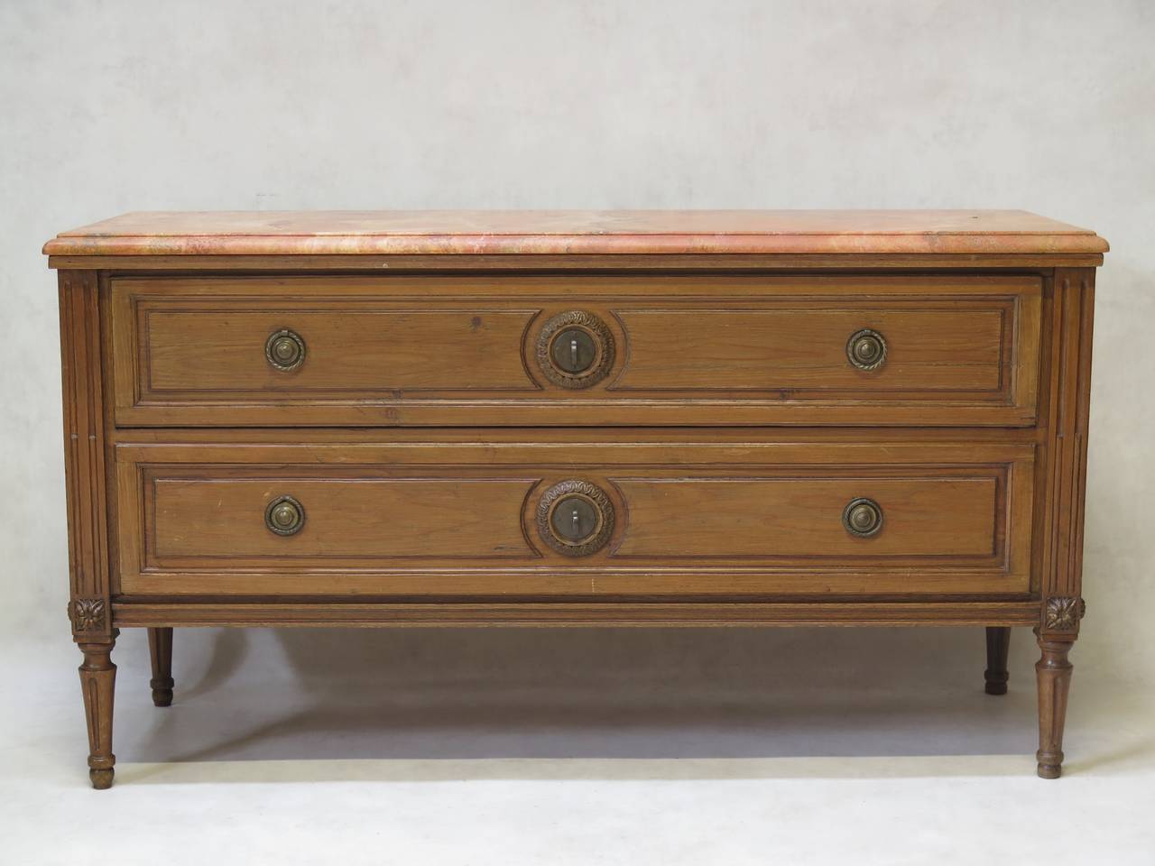 Charming Louis XVI style two-drawer commode in natural waxed pine wood, with a pink travertine top. Raised on tapering and fluted legs. Carved oval medallion detail on either side. The long drawers were originally destined to hold trousers hence the
