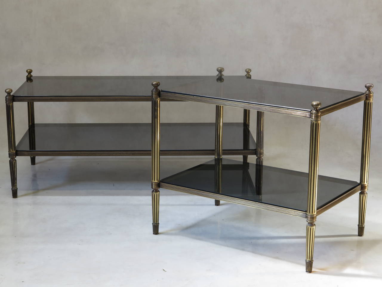 Very chic and elegant almost-pair of solid brass (or bronze) coffee tables, with smoked glass tops.

The table tops have canted corners. The top tiers are decorated at each corner with an engraved finial. Reeded legs, ending in elegant feet. The