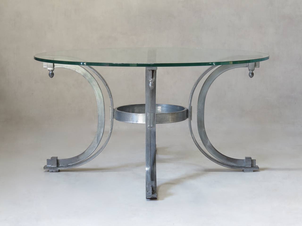 Chic neoclassical style round coffee table. The thick clear glass top is supported by four C-form legs ending in finials and joined by a circular stretcher.

We have four tables available. They are identical except for the finials, which vary from