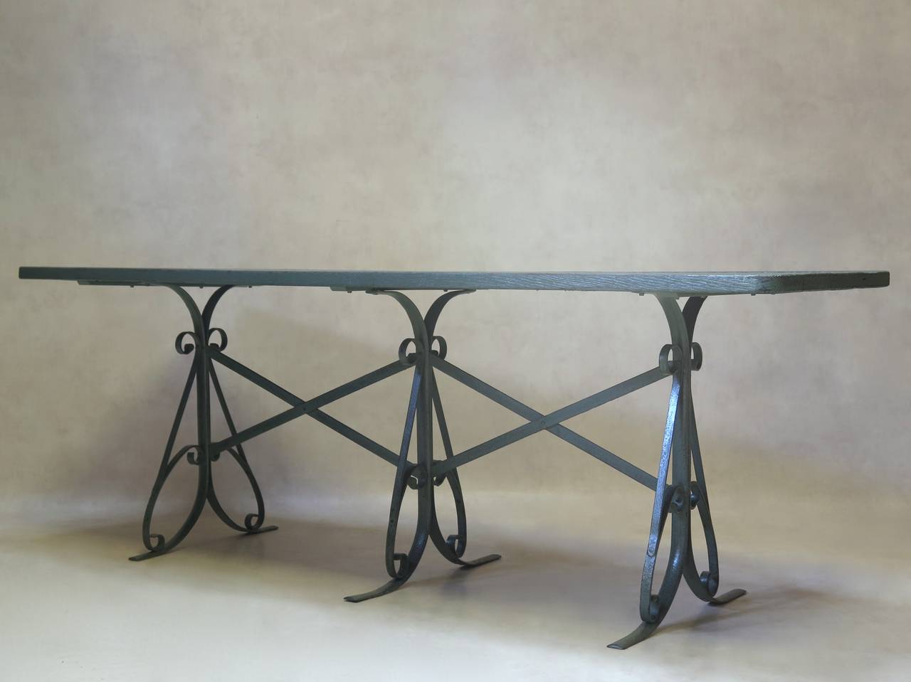 Dining table with a wrought-iron base and wood top, painted in soft grey-green. The three scrolling iron legs are joined together by X-shape stretchers. Can be used indoors or out.