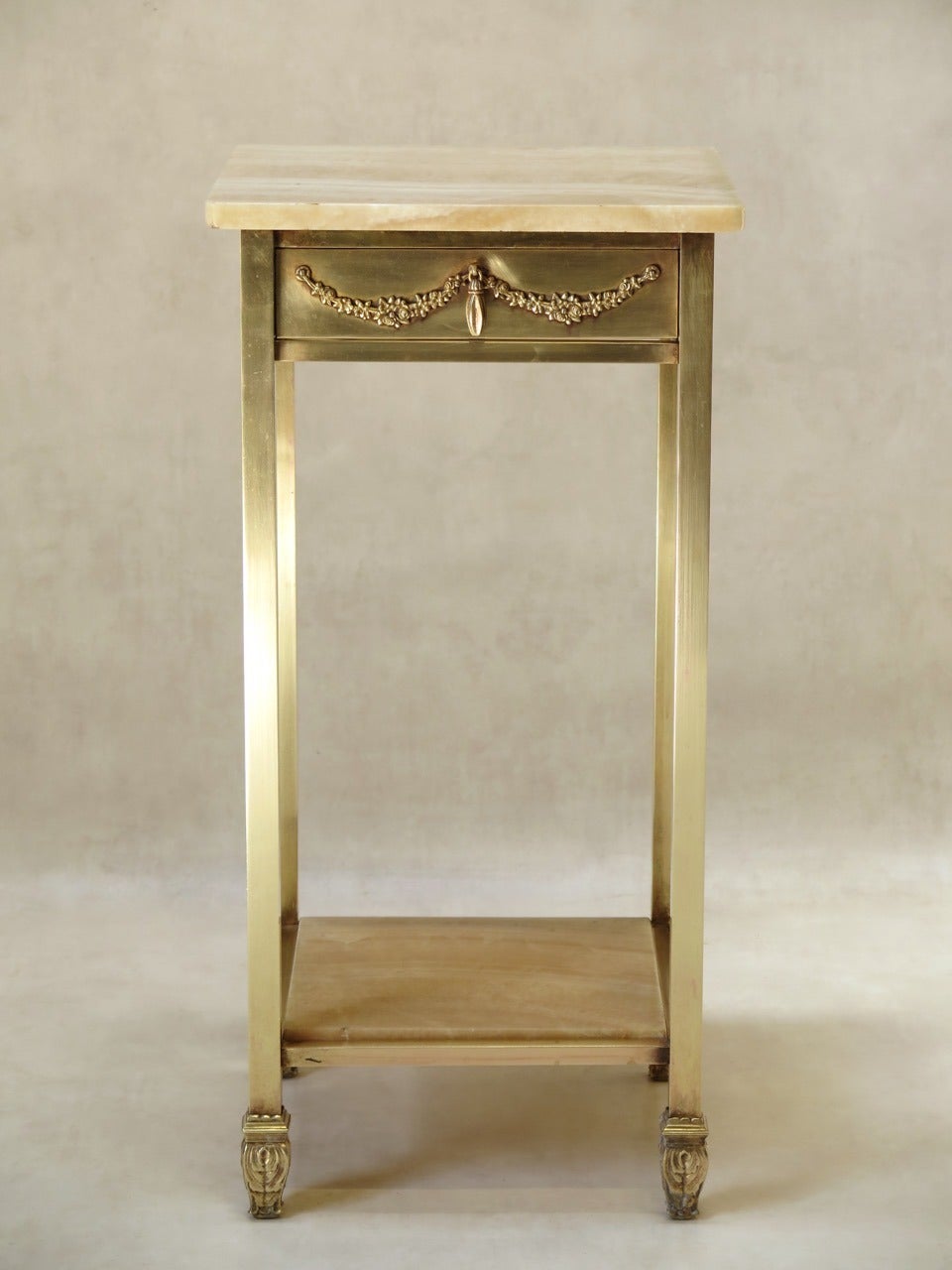 Chic night stand, with a solid brass structure and an onyx top and lower shelf. The drawer is embellished with a simple flower wreath. Lovely acanthus leaf feet. Heavy and well-made.