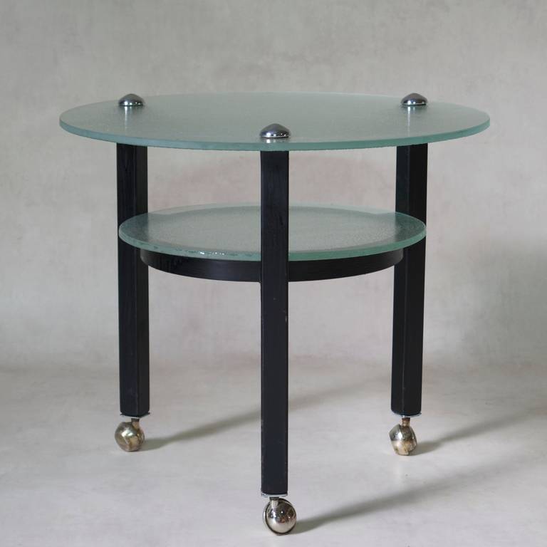 Table d'appoint chic et minimaliste avec deux étages de verre épais et texturé, et une structure métallique à trois pieds, peinte en noir, sur roulettes chromées.
