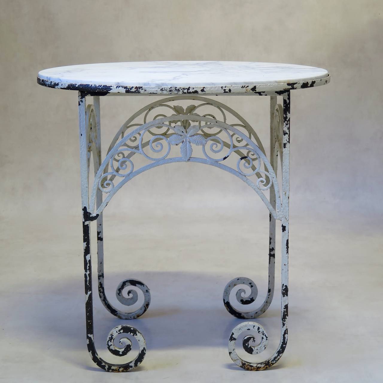 Delightful petite oval side table with a wrought iron base and a white marble top veined with grey. The base is unusual with its scrolled feet and pretty apron, decorated with leaves and a scrolling motif. Original, distressed, glossy white paint.
