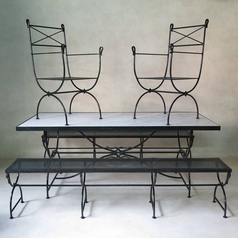A very chic 1940s wrought-iron outdoor dining-set, comprised of one long dining table with Carrarra marble top, two benches and two armchairs. The benches and chairs have cloverleaf-patterned sheet metal seats. 

Curule shape bases, with 