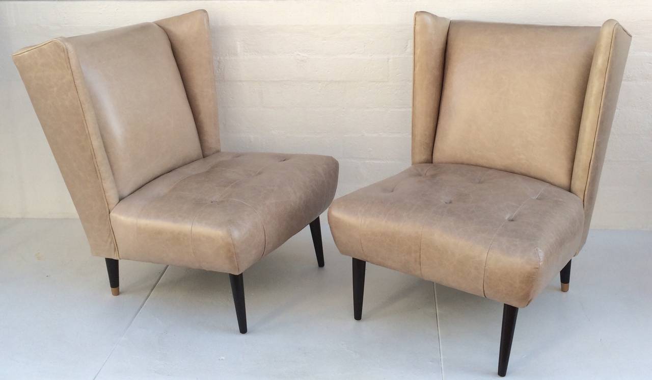 A pair of 1950s club chairs newly reupholster in a cream distressed leather, with wood tapered legs finished is a dark brown lacquer, with the back legs having brass caps.