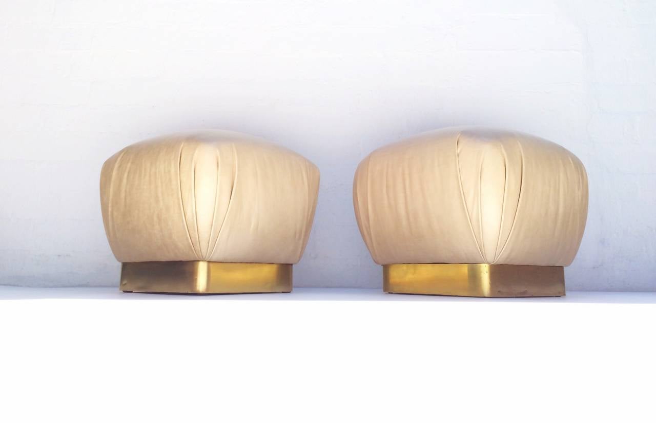 For your consideration, a pair of newly reupholstered leather poufs with polished brass bases. 
These poufs designed by Marge Carson and made in 1975 are reupholstered in a cream colored leather and the wood base frames are banded with polished