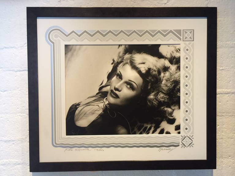 This is a signed black and white photograph of Rita Hayworth numbered 102/450 taken in 1944.
The serigraph art deco border was designed by Hurrell and is also signed.  
In a black wood frame with glass.