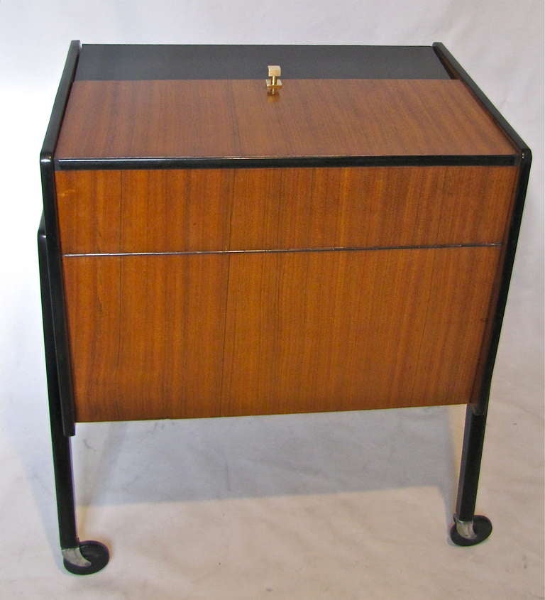Mid century modern wood bar cabinet in bleached walnut and ebony with molded rubber, resting on steel/plastic castors. Interior opens to bottle and cocktail glass storage.