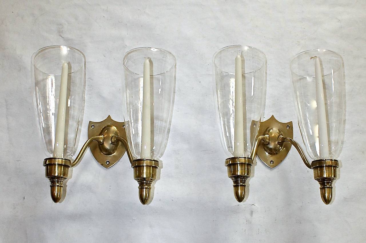 Pair of brass Federal style shield back 2 candle holder wall sconces with tall glass hurricane shades. Not electrified. Can be electrified upon request for a fee. a second pair that has been electrified is also available.