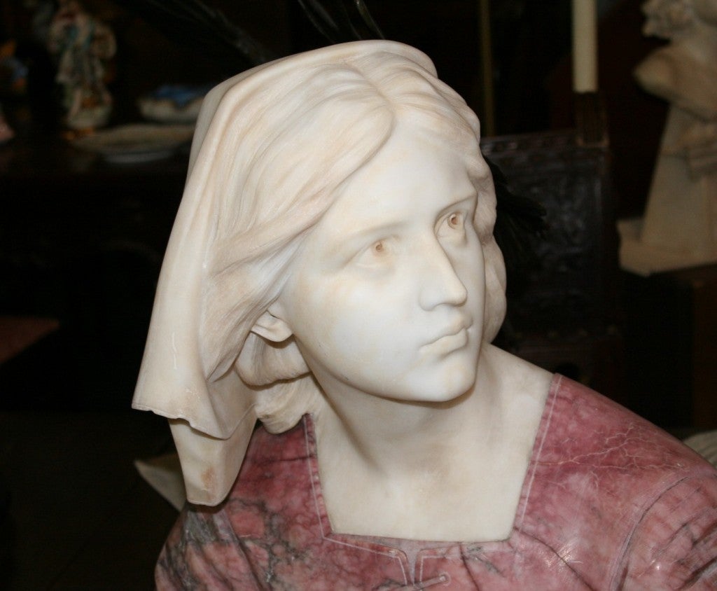 This is a hand carved bust of Saint Joan D'Arc. It is very well done by a listed sculptor of the 19th century. The shoulders are done in rouge alabaster and the head is carved from Italian Carrera Marble.