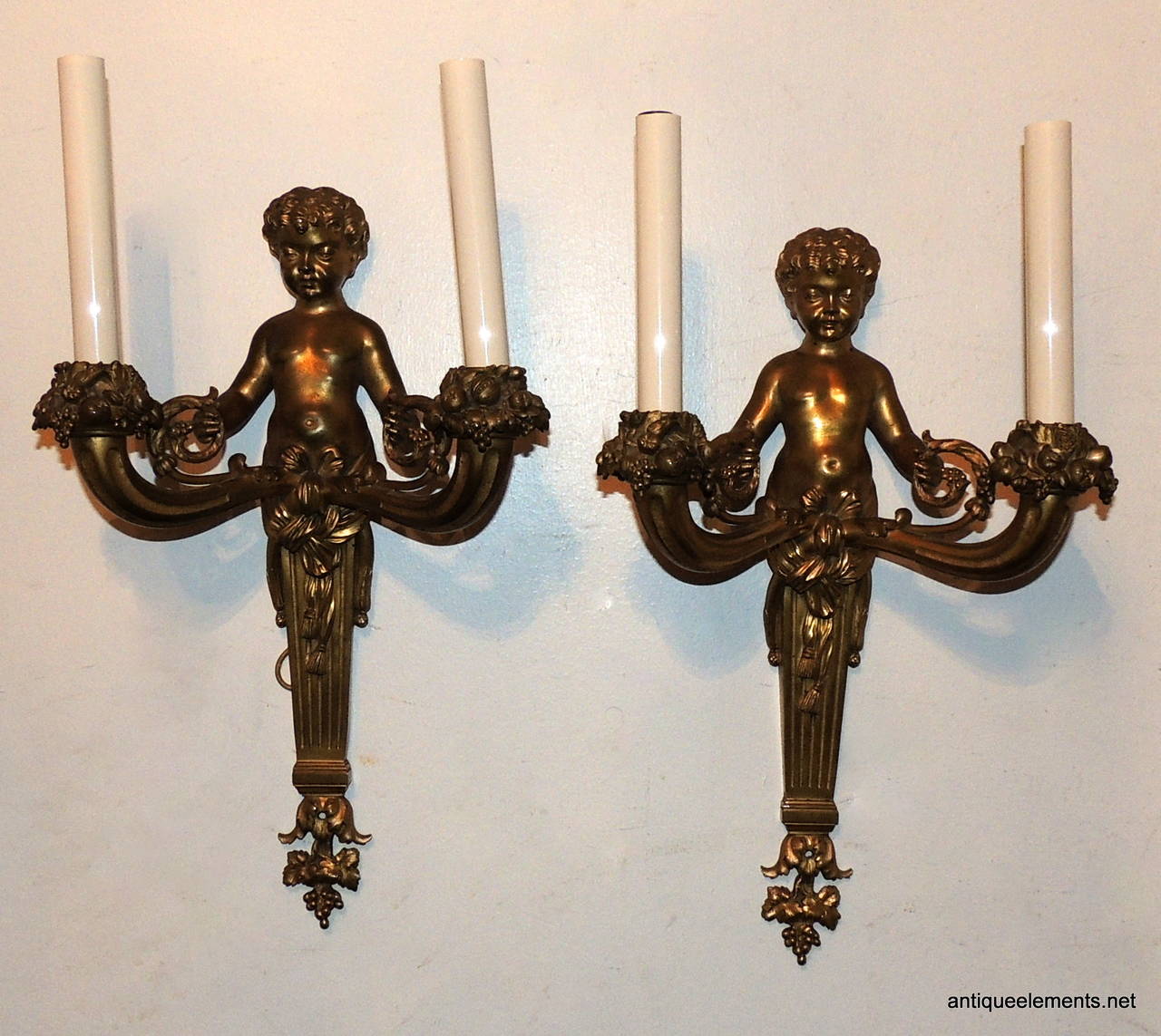 These wonderful French Doré bronze cherub two-arm sconces are full of detail. The bobeches are covered in floral and fruit draping and repeated at the end of the sconces. The bottom of the cherub has fabric draping and a bow that accents the center