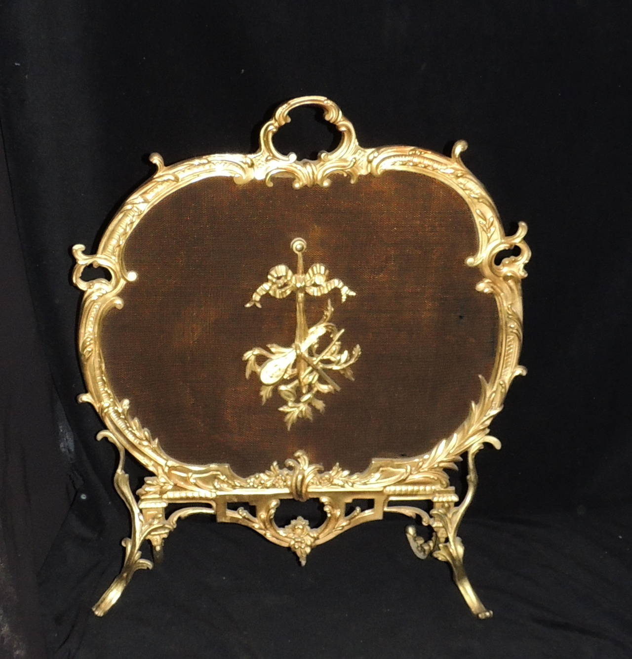 Elegante biombo rococó francés de bronce dorado decorado con ornamentos, empezando por el medallón central, que es un laúd con cuernos musicales atado con un bonito detalle de lazo. El asa y el trabajo en bronce que la rodea son bellos detalles de