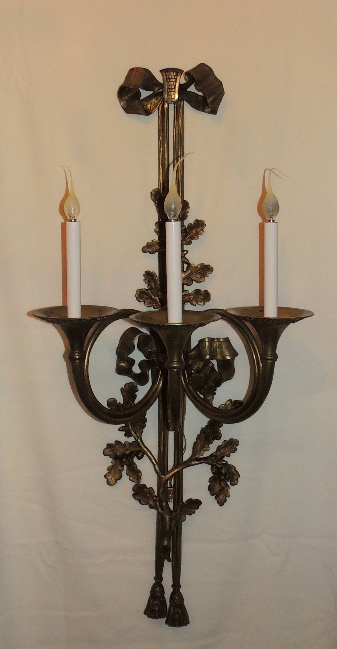 These wonderful pair of antique bronze three-arm sconces are three French horns holding the three lights. Starting at the top, these beautiful sconces are topped with a ribbon bow continuing with filigree details down the center and finished with