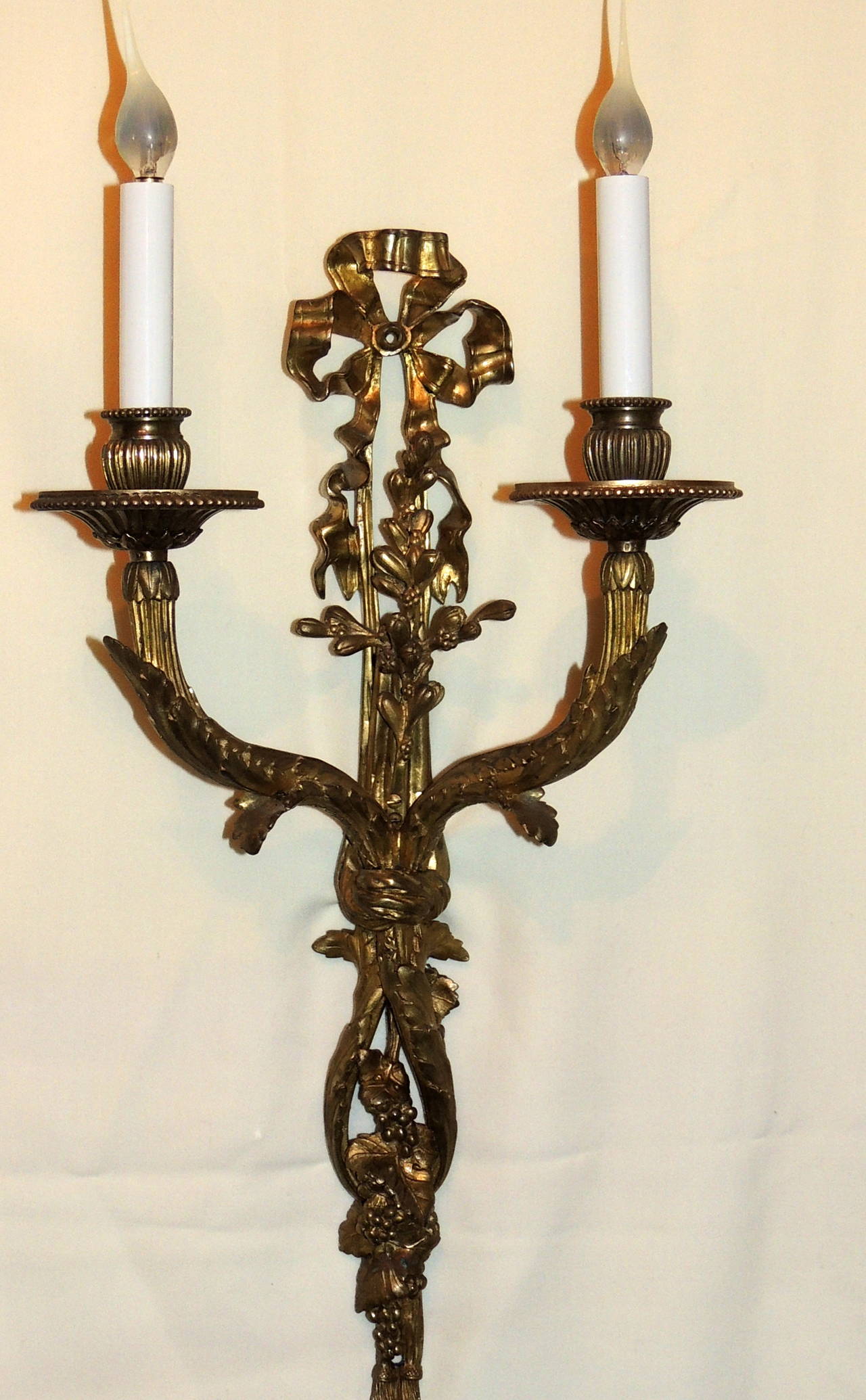 These elegant bow top sconces are early 20th century antique bronze with a wonderful aged patina. The bow with the cascading ribbons entwining the beautiful floral filigree and seed detail along both arms and the centre finish with the tassels at