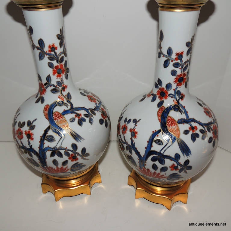 These beautifully hand-painted lamps with Asian influenced birds and beautiful floral detail are done in beautiful orange, red and blues with gilt detailing throughout. The neck and pedestal bottom are doré bronze with pierced detailing at the top