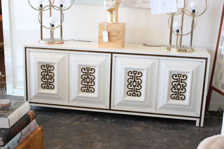 New lacquered Mastercraft credenza by William Doezema in ivory tusk white with decorative brass appliques on door fronts. Front of credenza has brass trim that surrounds the front surface.The doors on the left opens to reveal pull out drawers while