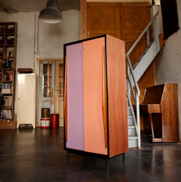 Sapeli tropic wood standing cabinet with relacquered multi -colored (violet-salmon pink) enamelled sliding doors.Black metal back.

The interior features 2 in height adjustable shelves and at the left, wardrobe with metal rod.Key is no longer
